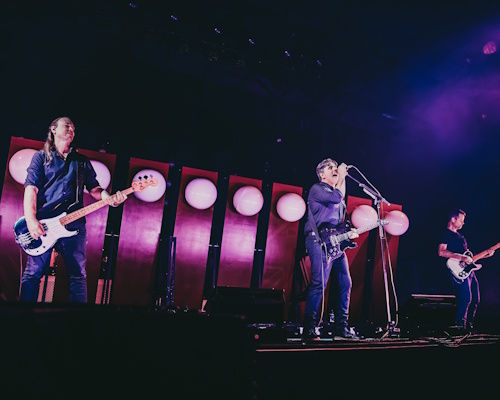 Three musicians perform on a purple-lit stage, singer at microphone center with guitars on either side and round lights behind.