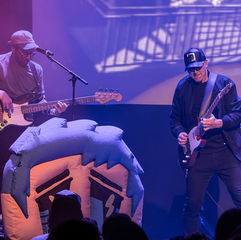 Two musicians perform on stage, one playing bass guitar and the other electric guitar. Stage is lit with purple lighting, backdrop visible.