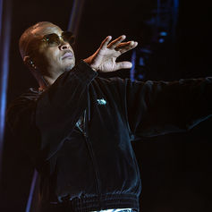 Man in sunglasses and a black jacket performs on a dark stage, gesturing with one hand while holding a microphone.