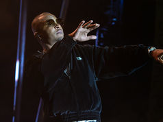 Man in sunglasses and a black jacket performs on a dark stage, gesturing with one hand while holding a microphone.