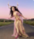 Woman with floral antlers in pink-gold gown kneels on a rural road at sunset. Deer in background, purple sky sets a mystical mood.
