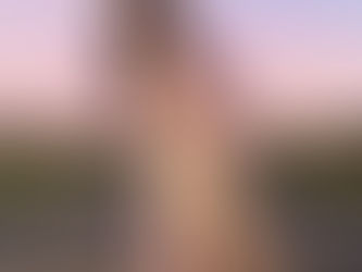 Woman with floral antlers in pink-gold gown kneels on a rural road at sunset. Deer in background, purple sky sets a mystical mood.