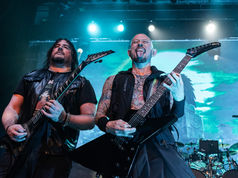 Two men playing electric guitars on stage, one sticking out his tongue. Blue lights and a screen with green patterns in the background. Rock vibe.