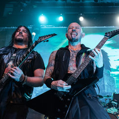 Two men playing electric guitars on stage, one sticking out his tongue. Blue lights and a screen with green patterns in the background. Rock vibe.