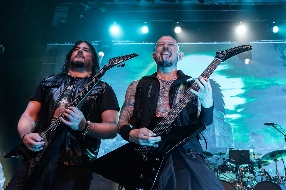 Two men playing electric guitars on stage, one sticking out his tongue. Blue lights and a screen with green patterns in the background. Rock vibe.