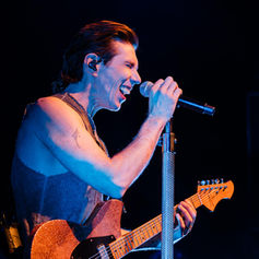 Josh Ramsay passionately performing on stage, holding a mic and playing a glittery electric guitar, with a deep blue-lit background.