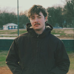 Young man with brown hair and mustache stands in a park wearing a dark jacket. Overcast sky, bare trees, and muted colors set a calm mood.