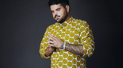 Man in a yellow spiral-patterned shirt with folded hands, looking contemplative. Neutral dark background, tattoo visible on forearm.