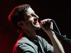 Smiling man sings into a microphone under bright stage lights, wearing a gray shirt. The background is dark with a hint of red lighting.