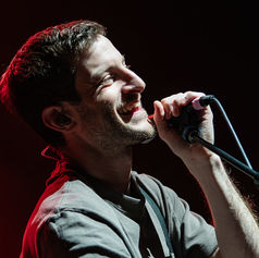 Smiling man sings into a microphone under bright stage lights, wearing a gray shirt. The background is dark with a hint of red lighting.