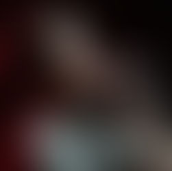 Smiling man sings into a microphone under bright stage lights, wearing a gray shirt. The background is dark with a hint of red lighting.