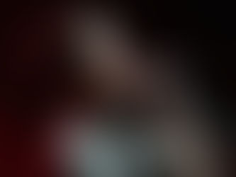 Smiling man sings into a microphone under bright stage lights, wearing a gray shirt. The background is dark with a hint of red lighting.