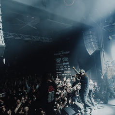 Band performing on stage at Manitoba Metalfest with a cheering crowd. Smoky atmosphere, speakers visible, and event posters in the background.