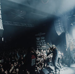 Band performing on stage at Manitoba Metalfest with a cheering crowd. Smoky atmosphere, speakers visible, and event posters in the background.