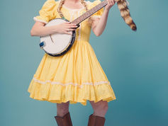 Person in yellow checkered dress playing a banjo, long blonde braids, and brown boots against a blue background, with a playful expression.