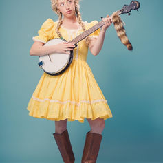 Person in yellow checkered dress playing a banjo, long blonde braids, and brown boots against a blue background, with a playful expression.