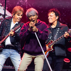 Three older rock musicians perform on a pink-lit stage, singer center with mic and two guitarists beside him, energetic and focused.