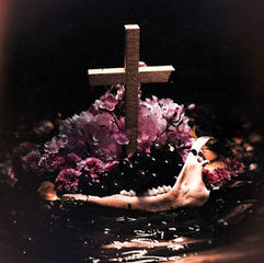 A wooden cross amid purple flowers and black grapes on water. A jawbone adds a mysterious, somber mood.