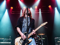 Rock guitarist sings into a mic on a smoky stage under red and blue lights, with Marshall amps and a drummer behind.