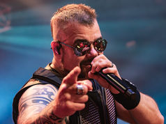 Singer in sunglasses points at the camera while singing into a microphone on a blue-lit stage.