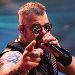 Singer in sunglasses points at the camera while singing into a microphone on a blue-lit stage.