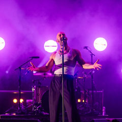 Singer passionately performing on stage with purple lighting. Spotlights and drums in background, creating a vibrant, energetic atmosphere.
