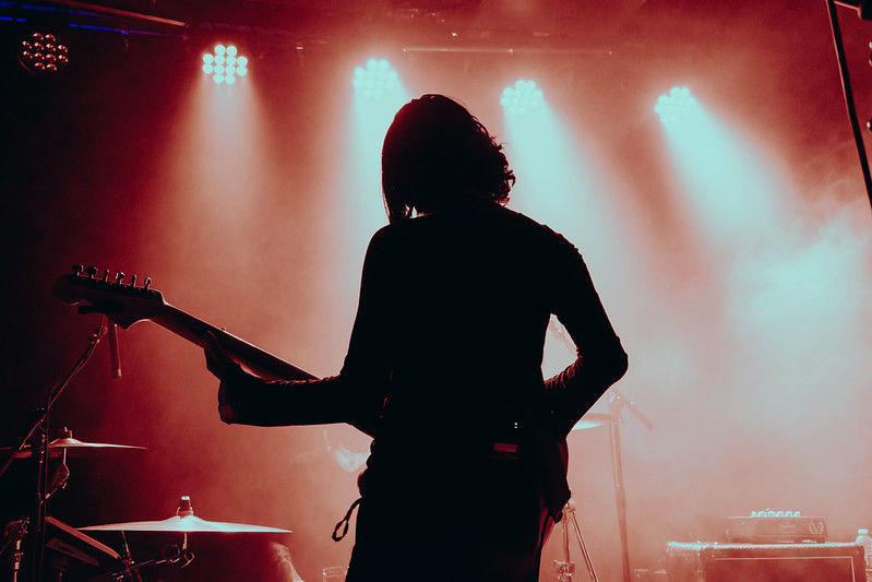 Silhouette of a guitarist on stage under red spotlights. Foggy atmosphere, drum set visible, creating a moody concert scene.