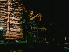 Musician on stage with guitar, forming a heart with hands. Dim lighting, textured backdrop, warm tones create an intimate concert vibe.