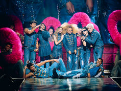 Smiling dance troupe in blue denim poses onstage amid giant pink fuzzy arches under bright concert lights.
