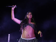 Jessie Reyez joyfully holding microphone on stage, wearing polka-dot outfit. Dark background with spotlight, creating an energetic, vibrant atmosphere.