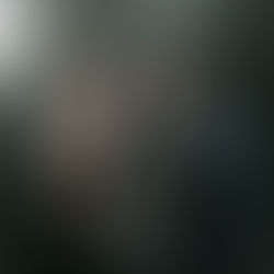 Man and woman seated facing each other in a dimly lit studio. Woman looks upwards; man is pensive. Dark clothes, neutral background.