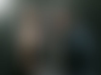 Man and woman seated facing each other in a dimly lit studio. Woman looks upwards; man is pensive. Dark clothes, neutral background.