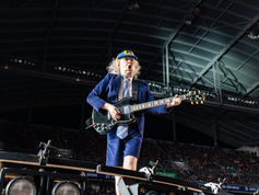 Angus Young in blue suit and cap plays black electric guitar on a stadium stage, with a crowd and roof lights behind him.
