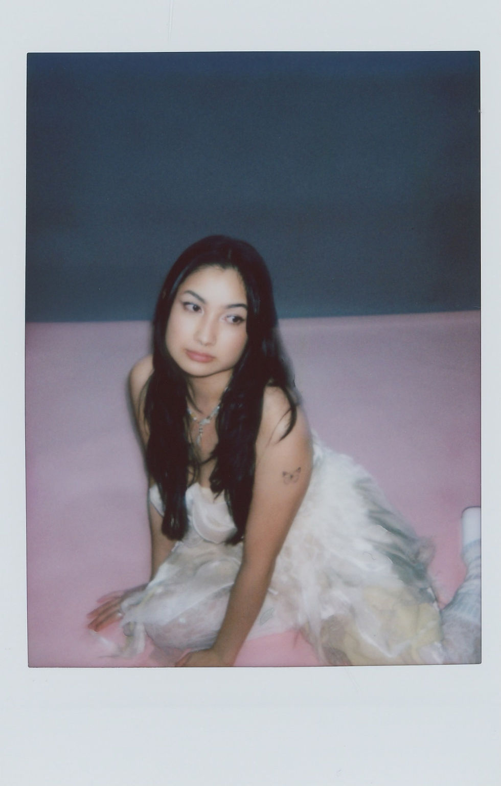 Glenna Jane in a flowing white dress kneels on a pink floor, gazing sideways. Gray background, butterfly tattoo on arm, calm expression.