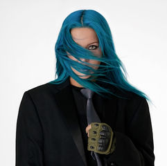 Charlotte Sands with blue hair in a suit, tie, and tactical glove, standing in front of a white background. Their expression is confident.