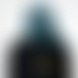 Charlotte Sands with blue hair in a suit, tie, and tactical glove, standing in front of a white background. Their expression is confident.