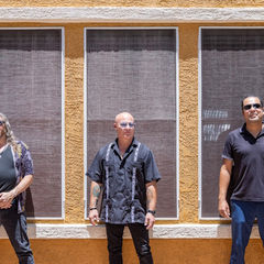 Three men stand confidently against an orange textured wall with three windows. They wear sunglasses, casual shirts, and exude a relaxed mood.