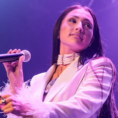 Female Indigenous singer in a white feather-trim jacket holds a microphone on a purple-lit stage, gazing off to the side.