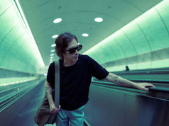 Person with sunglasses on an escalator in a modern tunnel, touching the railing. The setting has a teal hue, creating a calm mood.