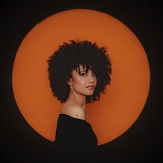 Milena Casado with curly hair in black top against an orange circle backdrop, looking over her shoulder with a serene expression.