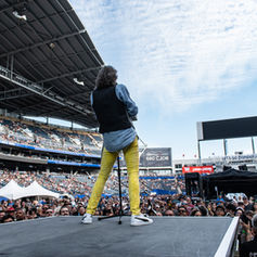 Kelly Hansen in yellow pants performing on stage to a large, cheering crowd in a stadium. Overhead roof and "Let's Go Bombers" banner visible.