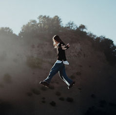 Person mid-jump in black top and jeans against a rocky hillside under a pale sky, with a dreamy mood.