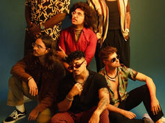 The six members of The Free Label pose in stylish, colorful outfits against a blueish green backdrop. They exude a cool, confident vibe, with relaxed expressions and sunglasses.
