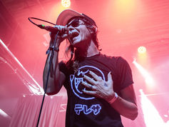 Singer in sunglasses and cap belts into a microphone on a pink-lit stage, wearing a black neon logo T-shirt with Japanese text.