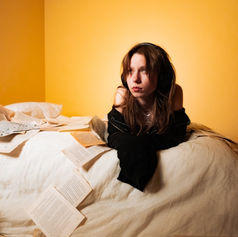 Teen girl with headphones and a black jacket sits on a bed in a yellow room, surrounded by scattered pages, looking contemplative.