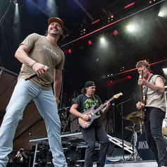 Band performing on an outdoor stage, smiling singer with CHENE shirt and guitarists under bright concert lights, energetic scene