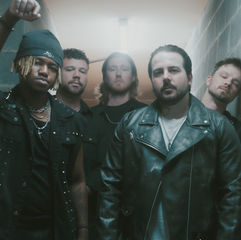 Five men in black clothing pose in a narrow hallway, one raising a fist, with a moody, smoky green-gray backdrop.