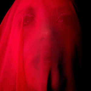 Face obscured by red fabric, creating a moody, mysterious vibe. Background is dark, enhancing the vivid red draping over the features.