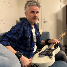 Man with grey hair and blue plaid shirt holds a white guitar on a blue couch in a room with a white door and wall covered in signatures.