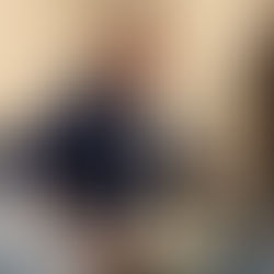 Man with grey hair and blue plaid shirt holds a white guitar on a blue couch in a room with a white door and wall covered in signatures.